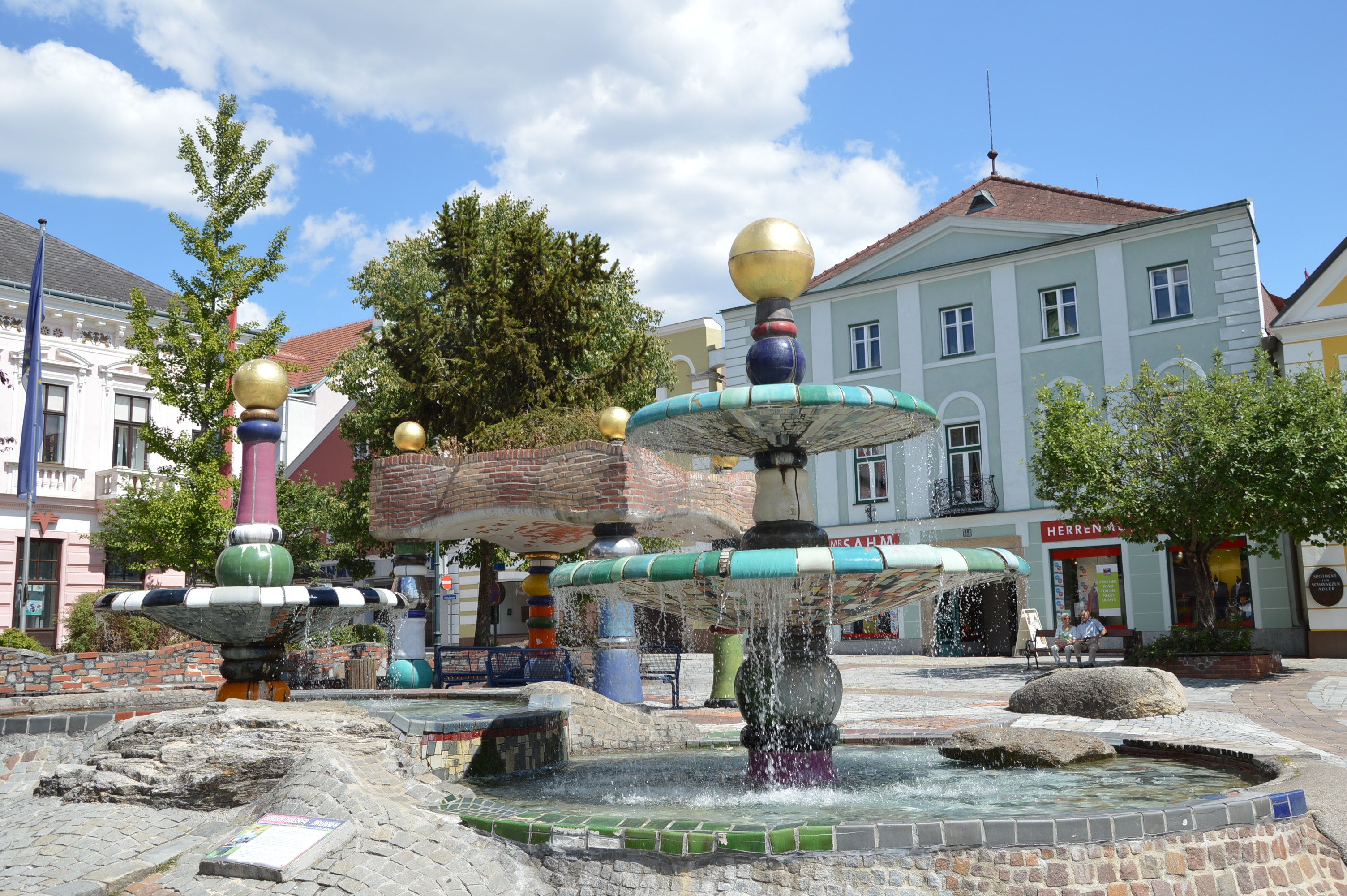 Barevná fontána ve stylu Hundertwassera se zlatými koulemi a tekoucí vodou, obklopená budovami a stromy.
