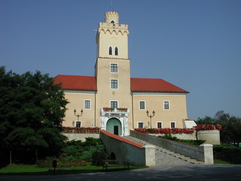Hrad s věží a červenými střechami, obklopený květinami a stromy, pod jasně modrou oblohou.