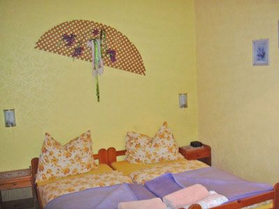 Útulná ložnice se žlutými stěnami, manželskou postelí se vzorovanými polštáři a dekorativním nástěnným ventilátorem.