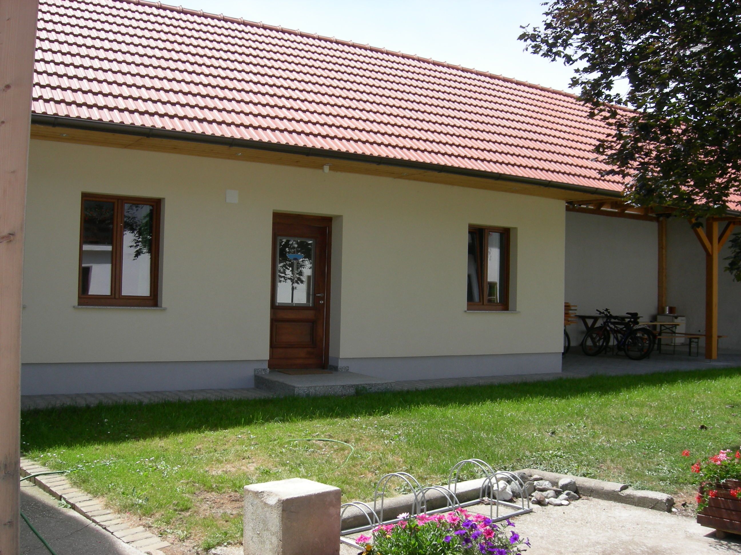 Vnitřní dvůr s budovou s červenou taškovou střechou, dřevěnými dveřmi a okny. V popředí jsou vidět květiny a stojan na kola.