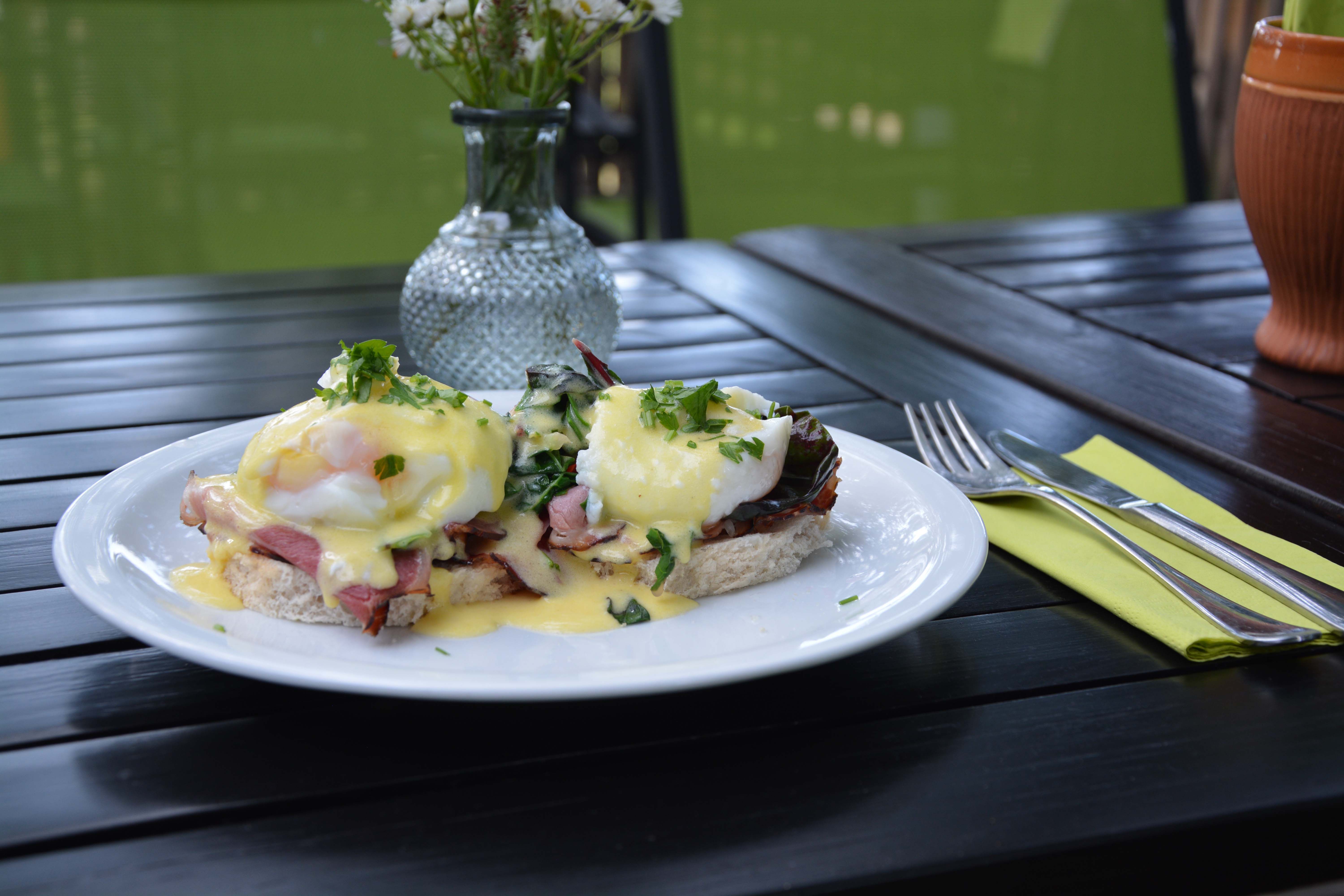 Talíř s vejci Benedikt na dřevěném stole, vedle příbory a váza s květinami.