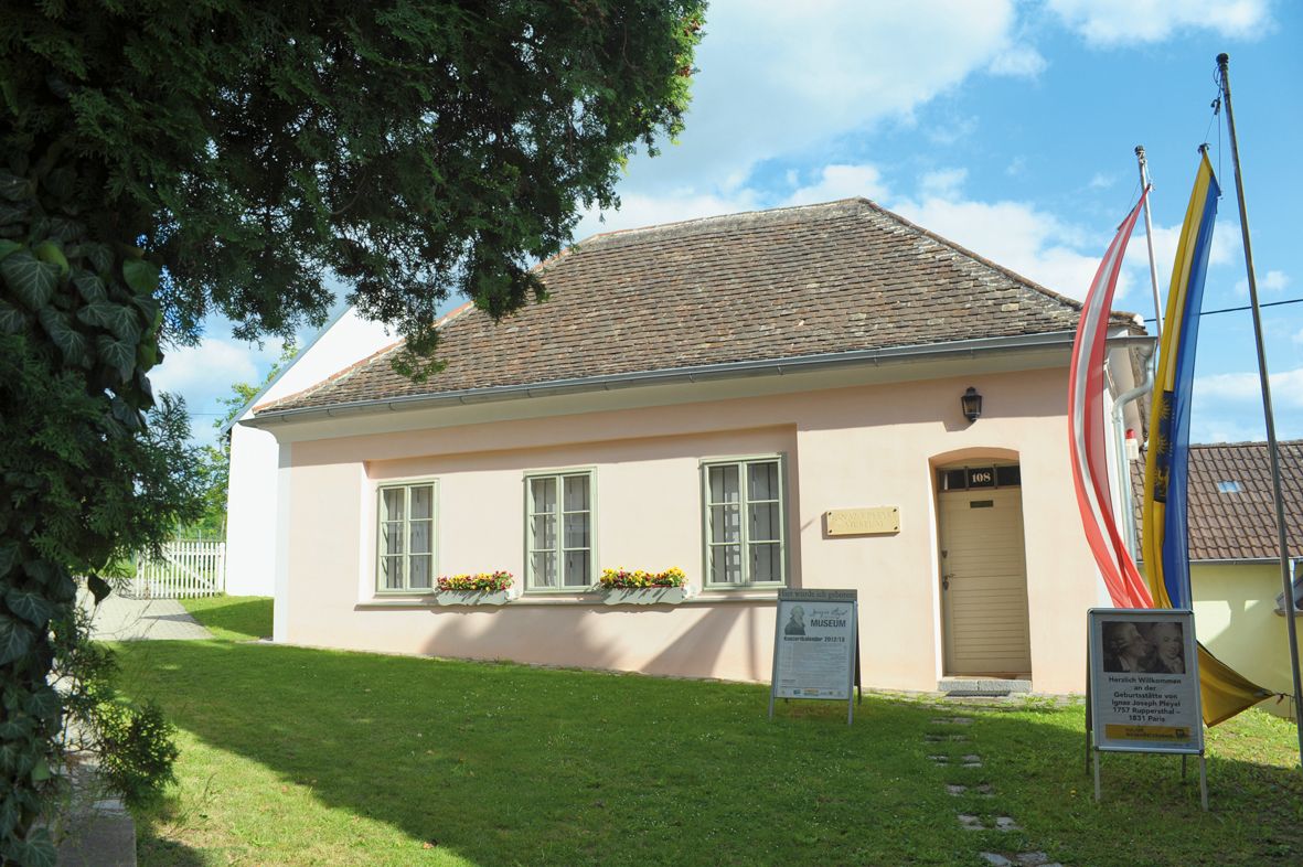 Malá, růžově natřená budova se šindelovou střechou, obklopená stromy a vlajkami, s cedulemi před vchodem.