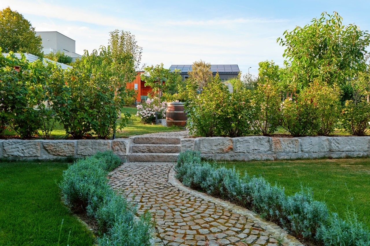 Udržovaná zahrada s kamennou cestou, keři a stromy, v pozadí budova se solárními panely.