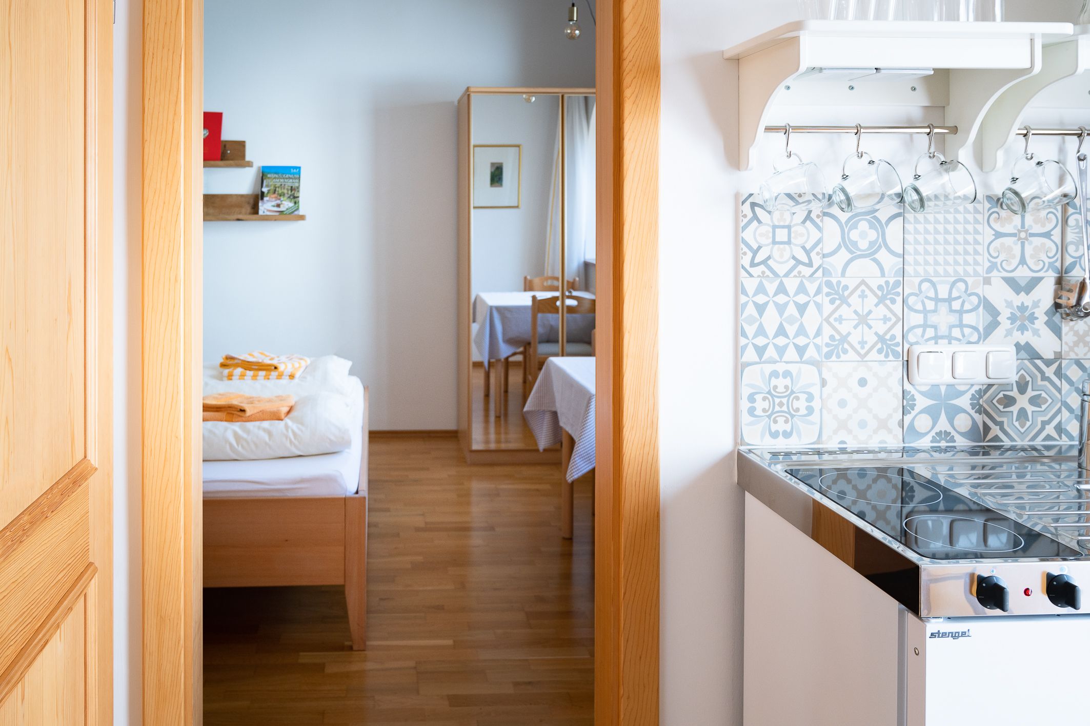 Pohled do interiéru pokoje s postelí, zrcadlem, stolem a malou kuchyňkou se vzorovanými dlaždicemi.