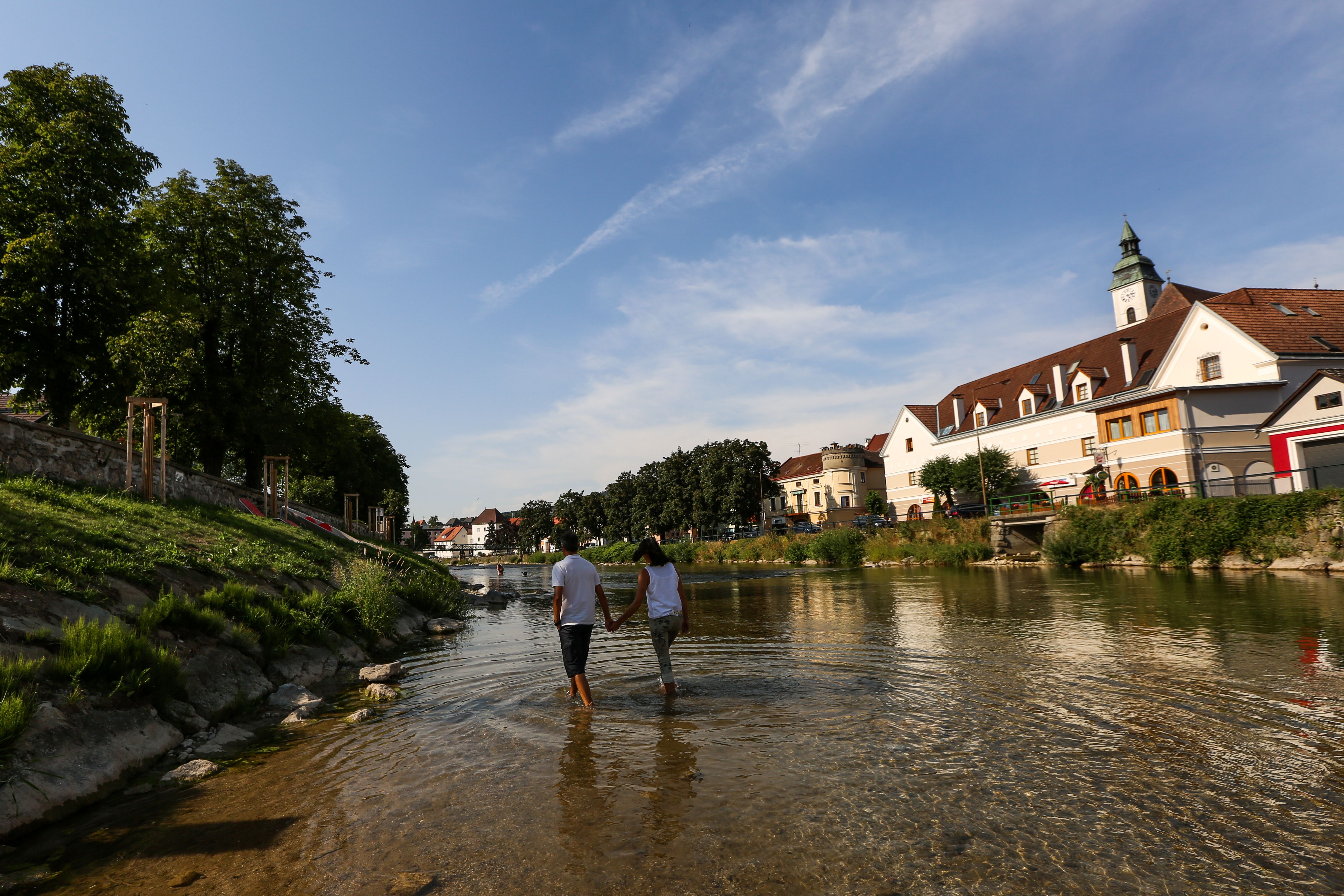 Pár se ruku v ruce prochází mělkou řekou ve městě s historickými budovami a stromy.
