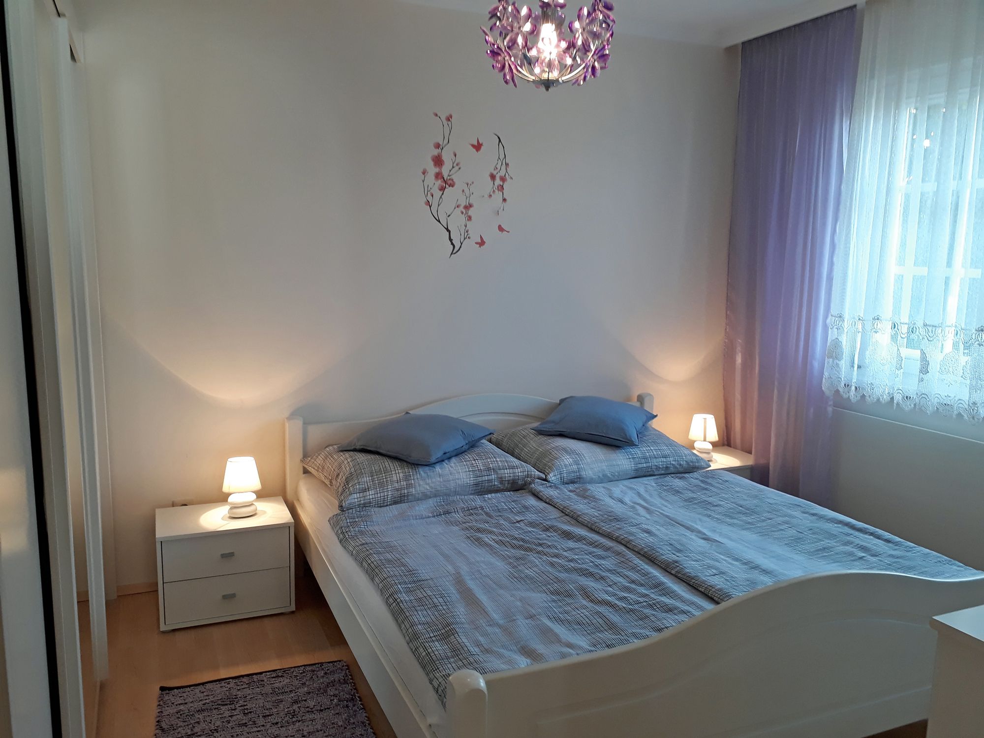 Útulná ložnice s manželskou postelí, dvěma nočními stolky s lampičkami a fialovými závěsy.