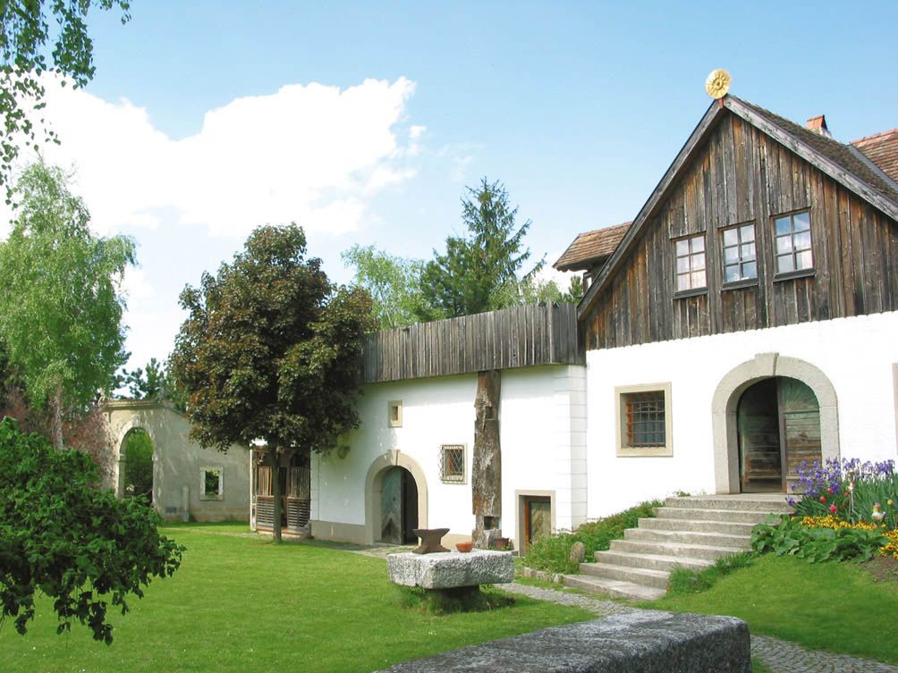 Tradiční budova s dřevěnou střechou a bílou fasádou, obklopená zelenou zahradou.