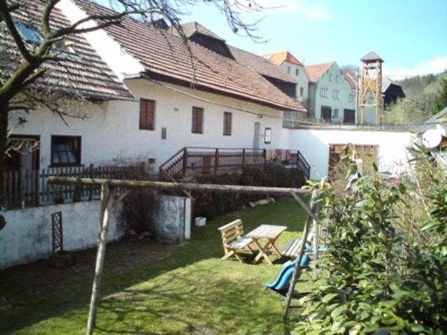 Venkovský dvůr s bílými budovami, dřevěným stolem a židlemi na zahradě, houpačkou a skluzavkou v popředí.