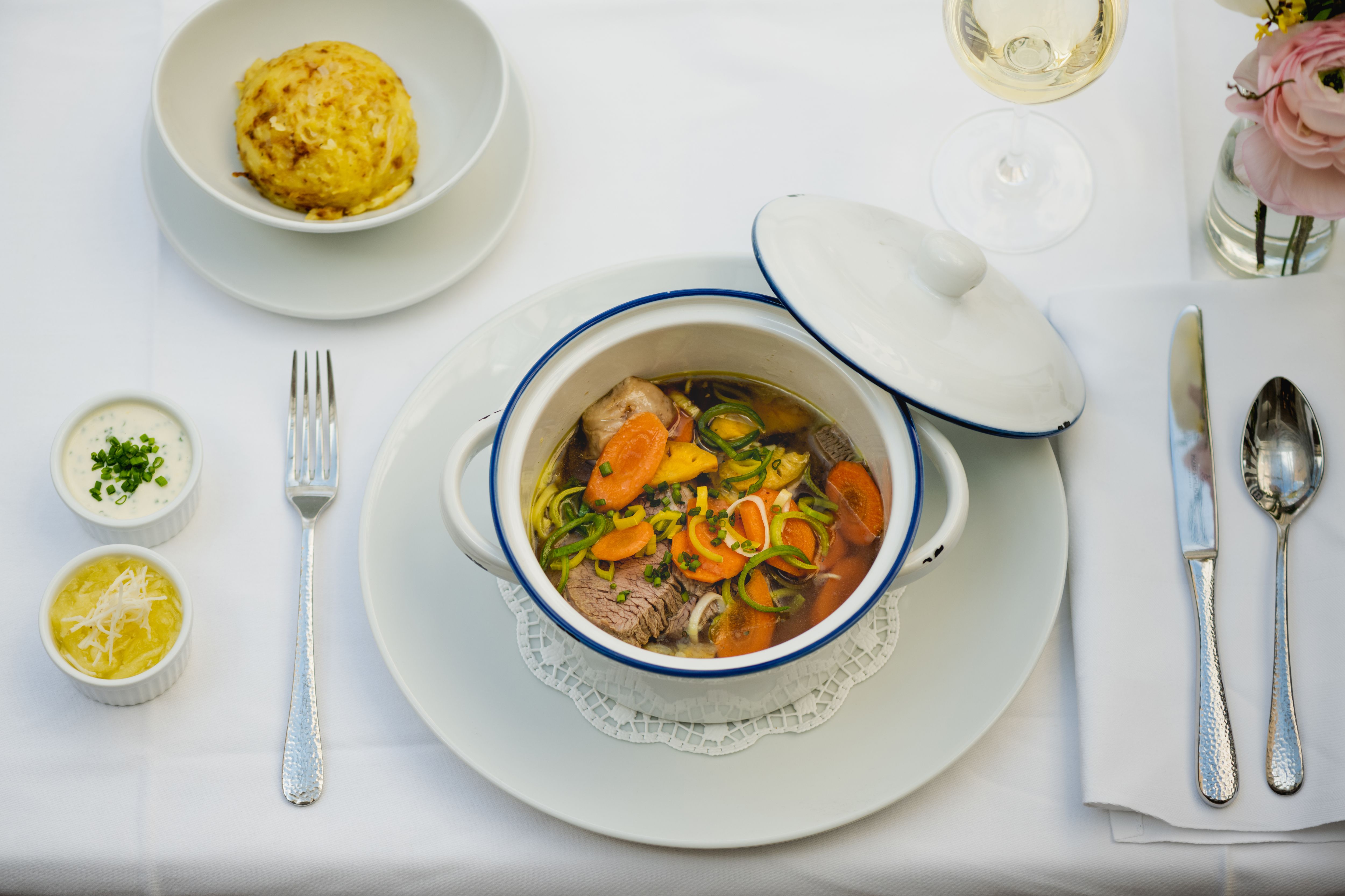 Stůl prostřený s gulášem v bílém hrnci, knedlíkem, dvěma omáčkami a sklenkou bílého vína.