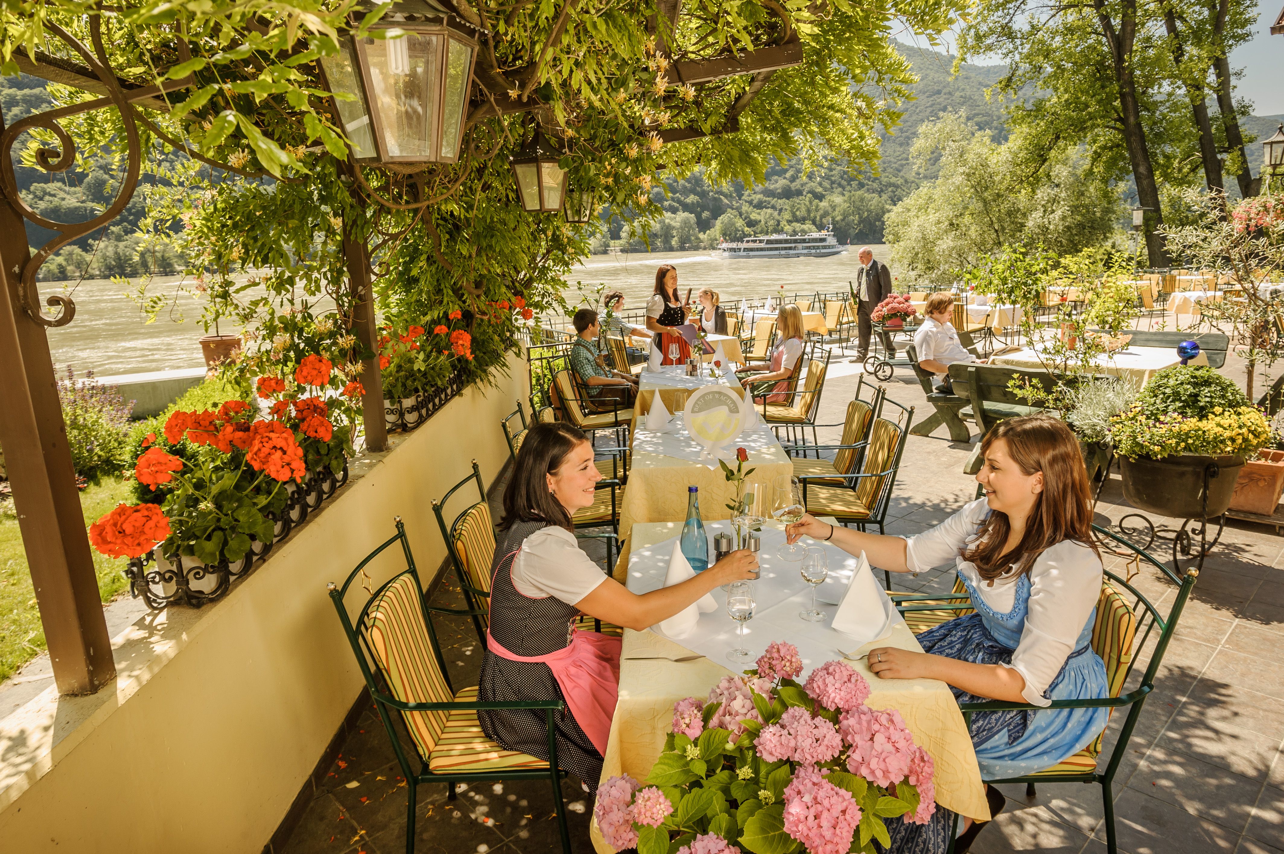 Lidé sedí v zahradní restauraci s výhledem na řeku, obklopeni květinami a stromy.