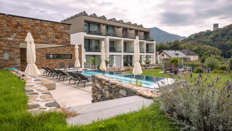 Pohled zvenčí na moderní butikový hotel s bazénem a lehátky, obklopený zelenou krajinou a kopci.
