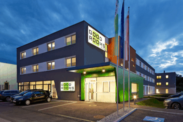 Moderní budova s nápisem "Good Rooms" za soumraku, obklopená auty a vlajkami.