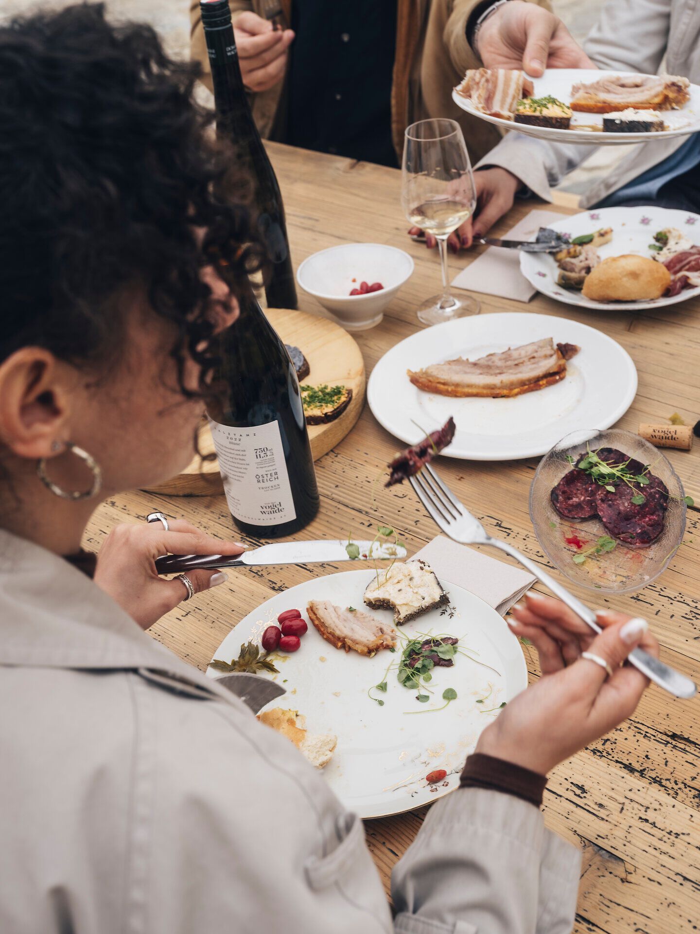 Mehrere Personen sitzen an einem Holztisch beim Heurigen; auf den Tellern liegen Brot, Aufstriche und kleine Speisen, dazu Gläser mit Wein.