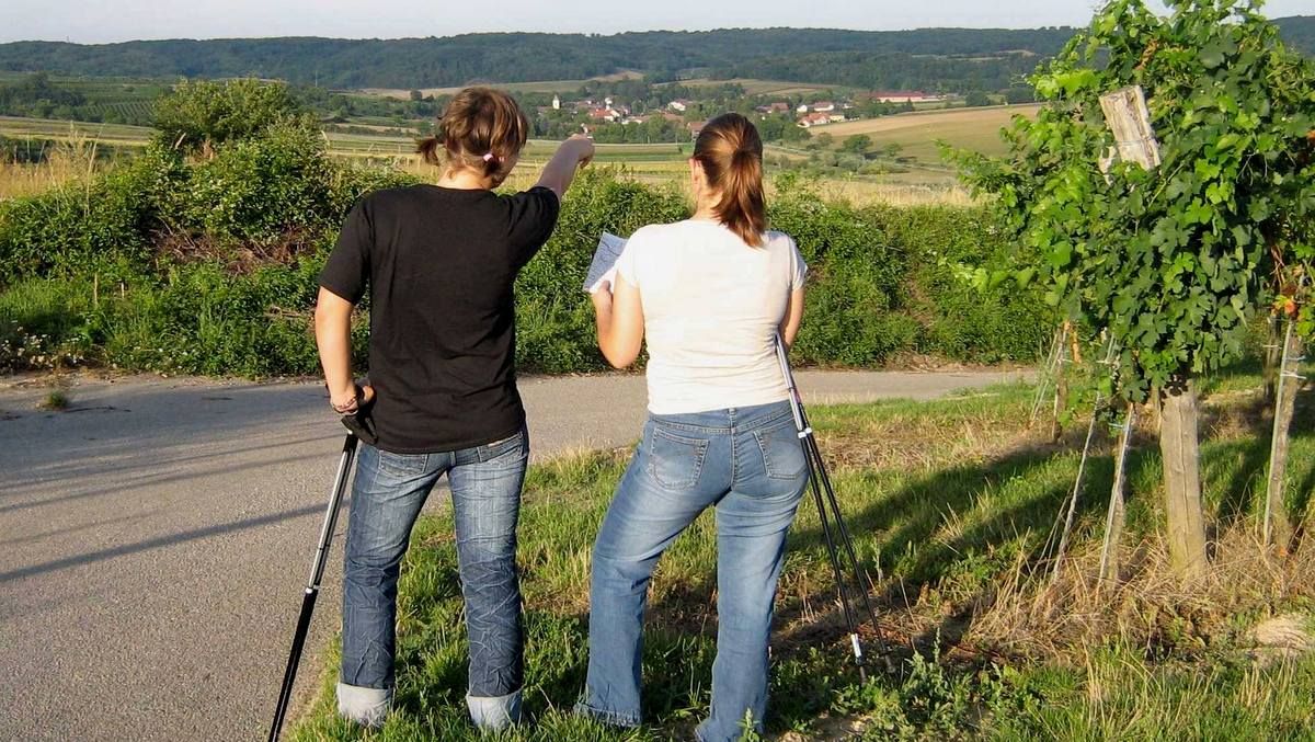 Dvě ženy s holemi stojí na cestě a dívají se do dálky, jedna ukazuje směrem k vesnici.