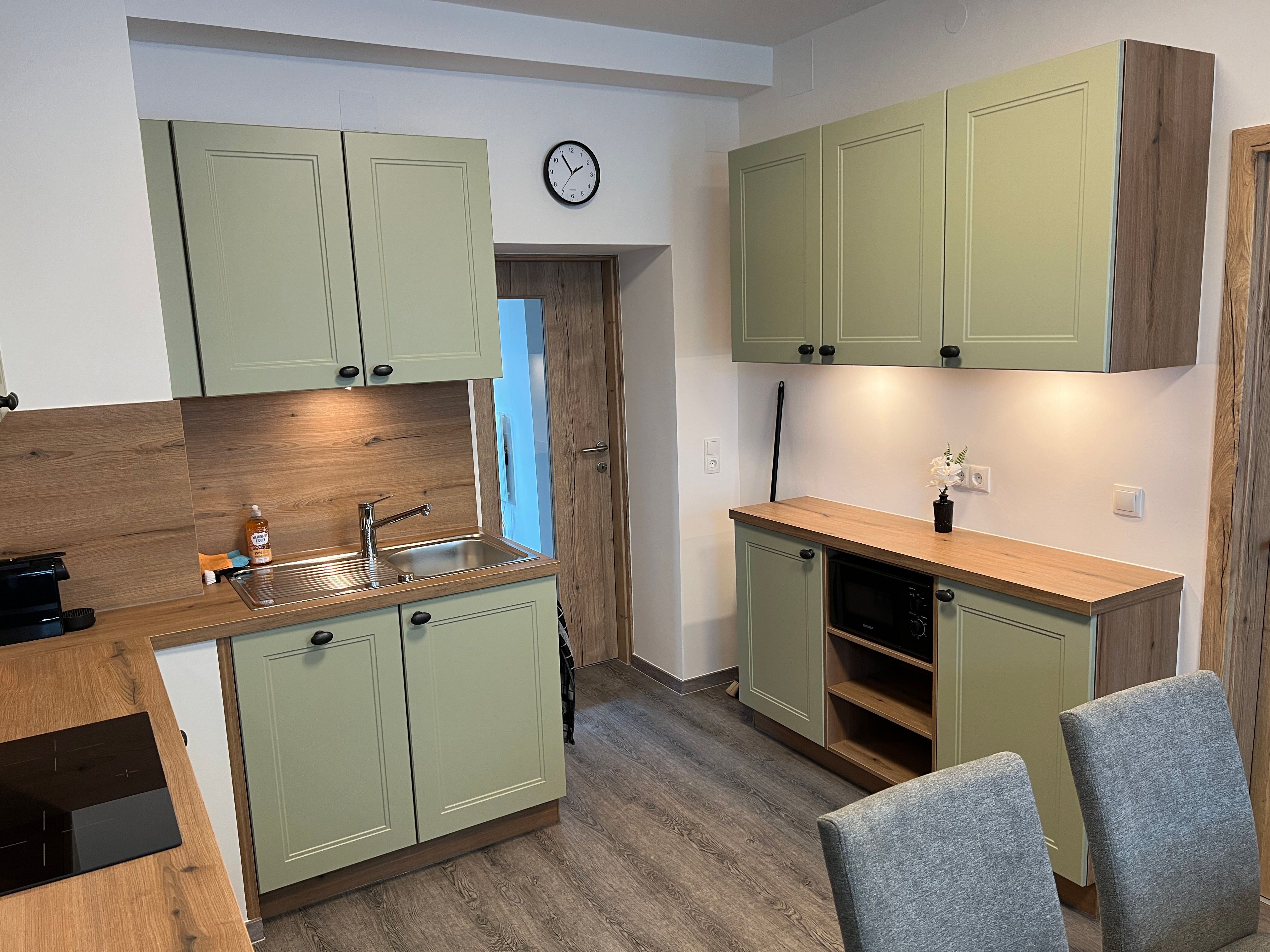 Moderní kuchyně se zelenými skříňkami, dřevěnou pracovní deskou a šedými židlemi.