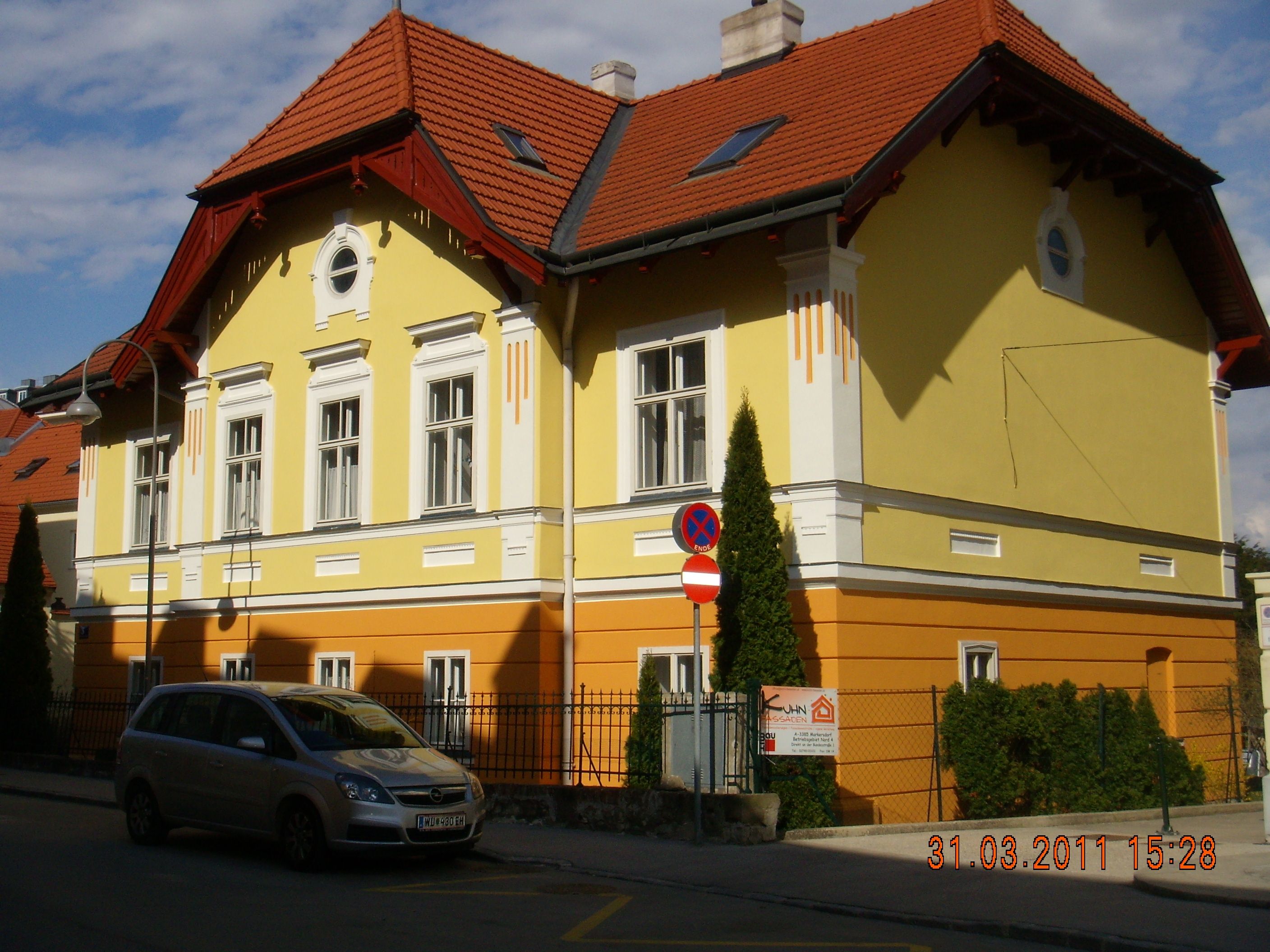 Žlutooranžová budova s červenou střechou a zaparkovaným autem před ní.
