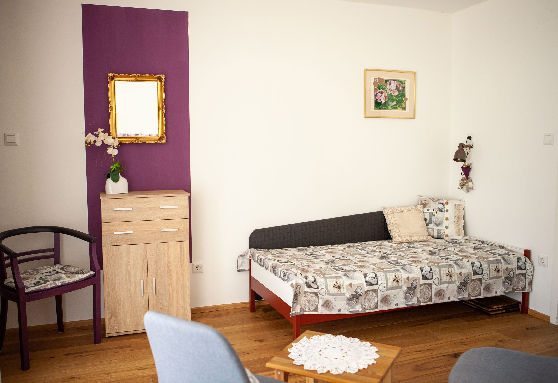 Obývací pokoj s rozkládací pohovkou, malou skříní, židlí a stolem na dřevěné podlaze.