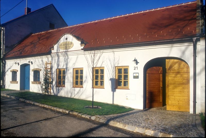Bílá budova s červenou střechou a dřevěnými dveřmi, označená "Kleinhaus".
