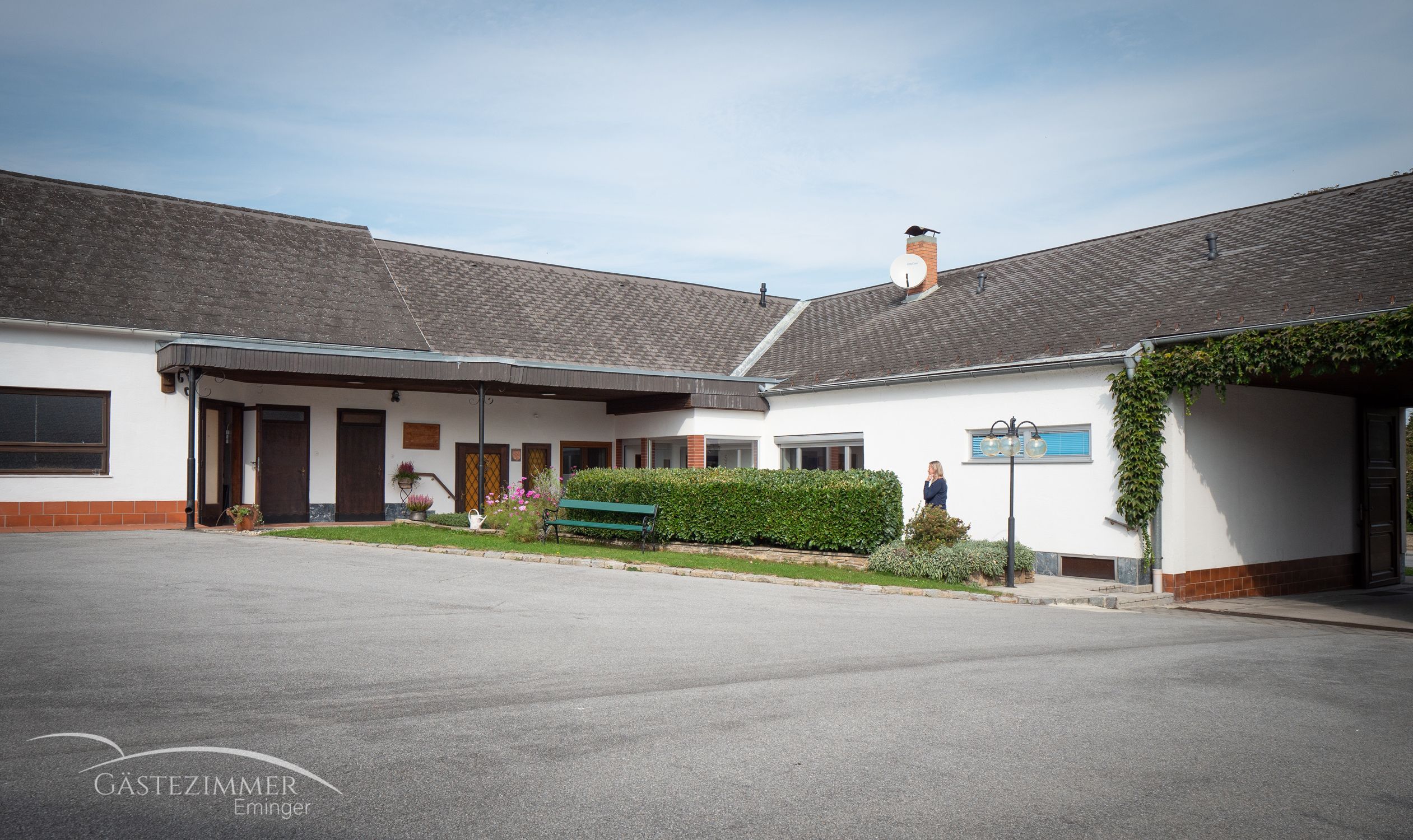 Dvůr s budovou, parkovacími místy a živým plotem. Vedle živého plotu stojí osoba. Na budově je vidět satelitní anténa.