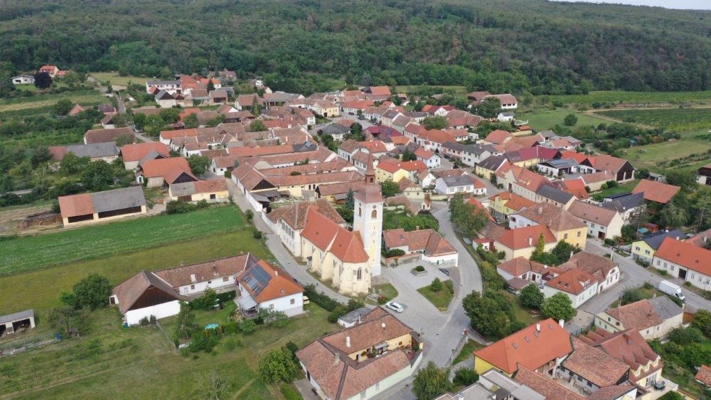 Letecký pohled na malou vesnici s kostelem v centru a červenými střechami, obklopenou zelenými poli a lesy.