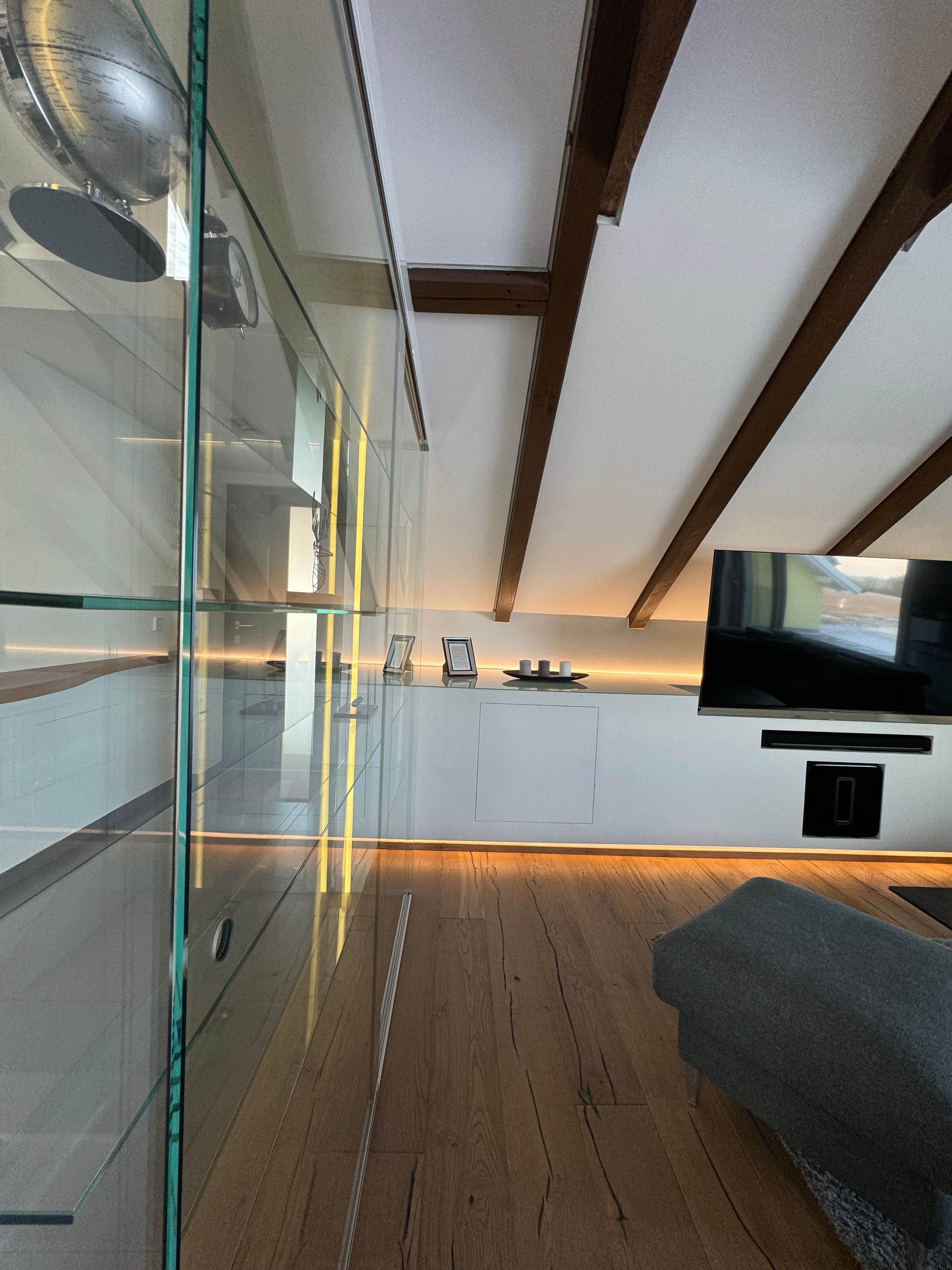 Moderní obývací pokoj s dřevěnou podlahou, prosklenou skříní, TV a šikmým stropem s dřevěnými trámy.