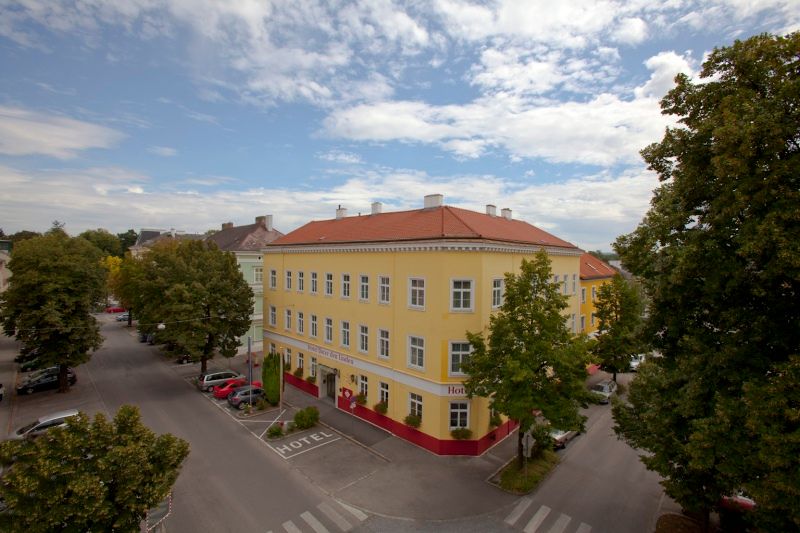 Pohled zvenčí na žlutý hotel s červenou střechou, obklopený stromy a ulicemi.