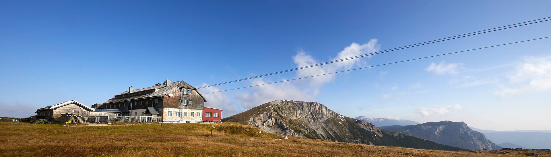 Panoramatický pohled na horskou chatu s okolními vrcholy pod modrou oblohou.