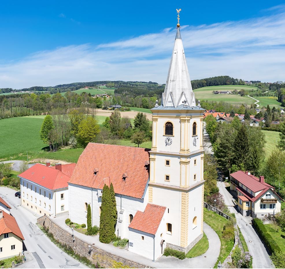 Letecký pohled na opevněný kostel v Krumbachu ve venkovském prostředí se zelenými poli a modrou oblohou.