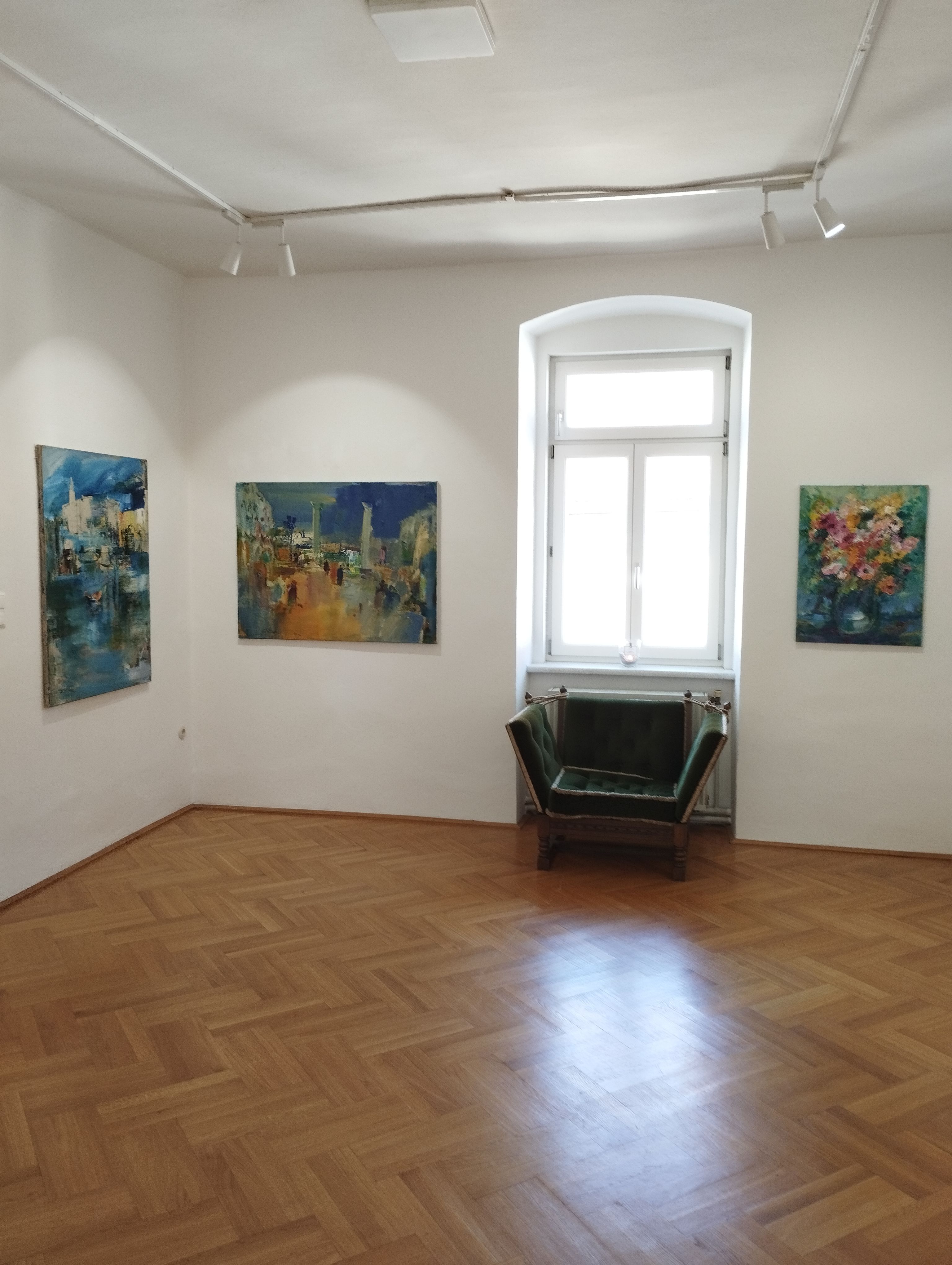 Pohled do interiéru galerie se třemi obrazy na stěnách a zeleným křeslem před oknem.