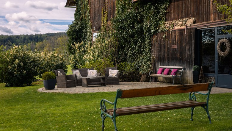 Zahrada s posezen&iacute;m před dřevěnou budovou porostlou břečťanem.