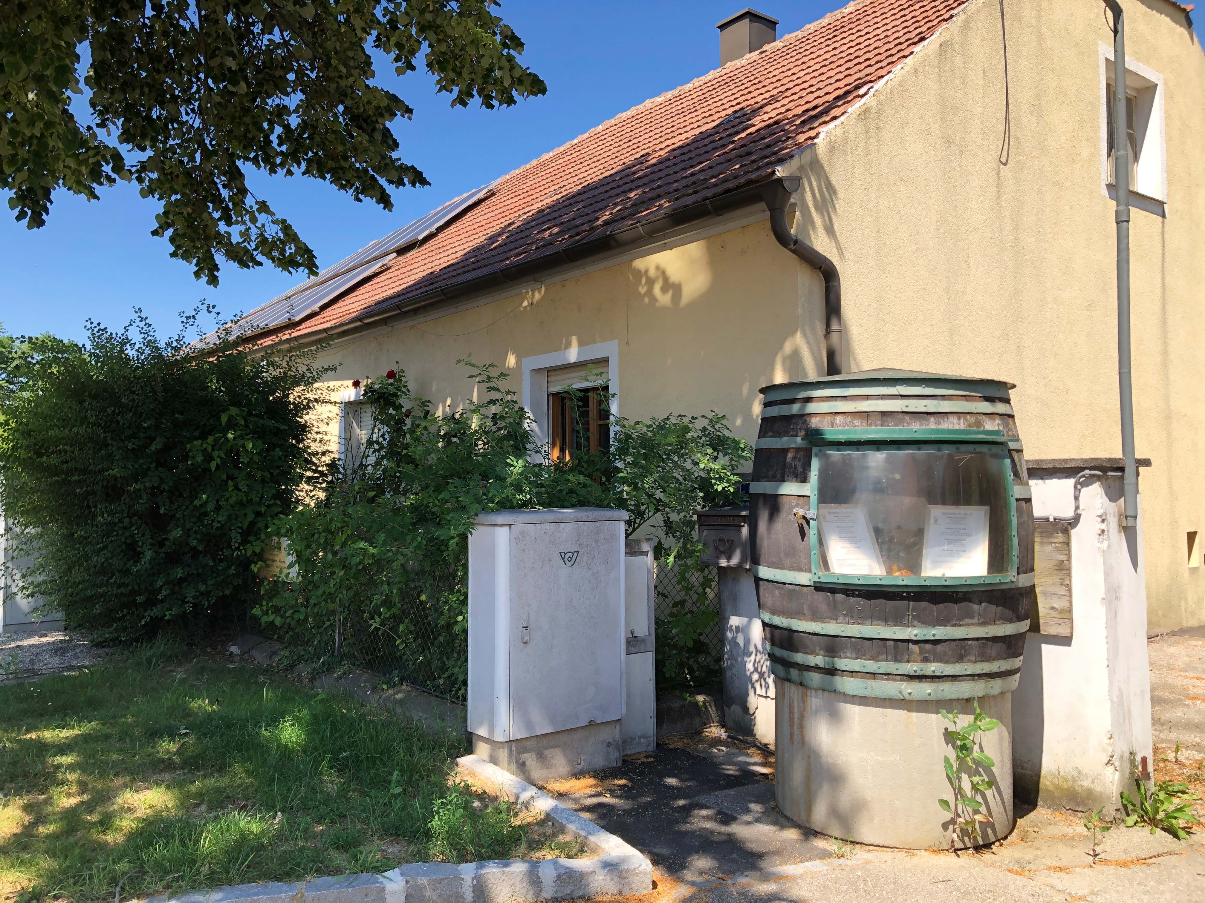 Žlutý dům s červenou taškovou střechou a solárními panely. V popředí velký sud s průzorem a rostlinami.