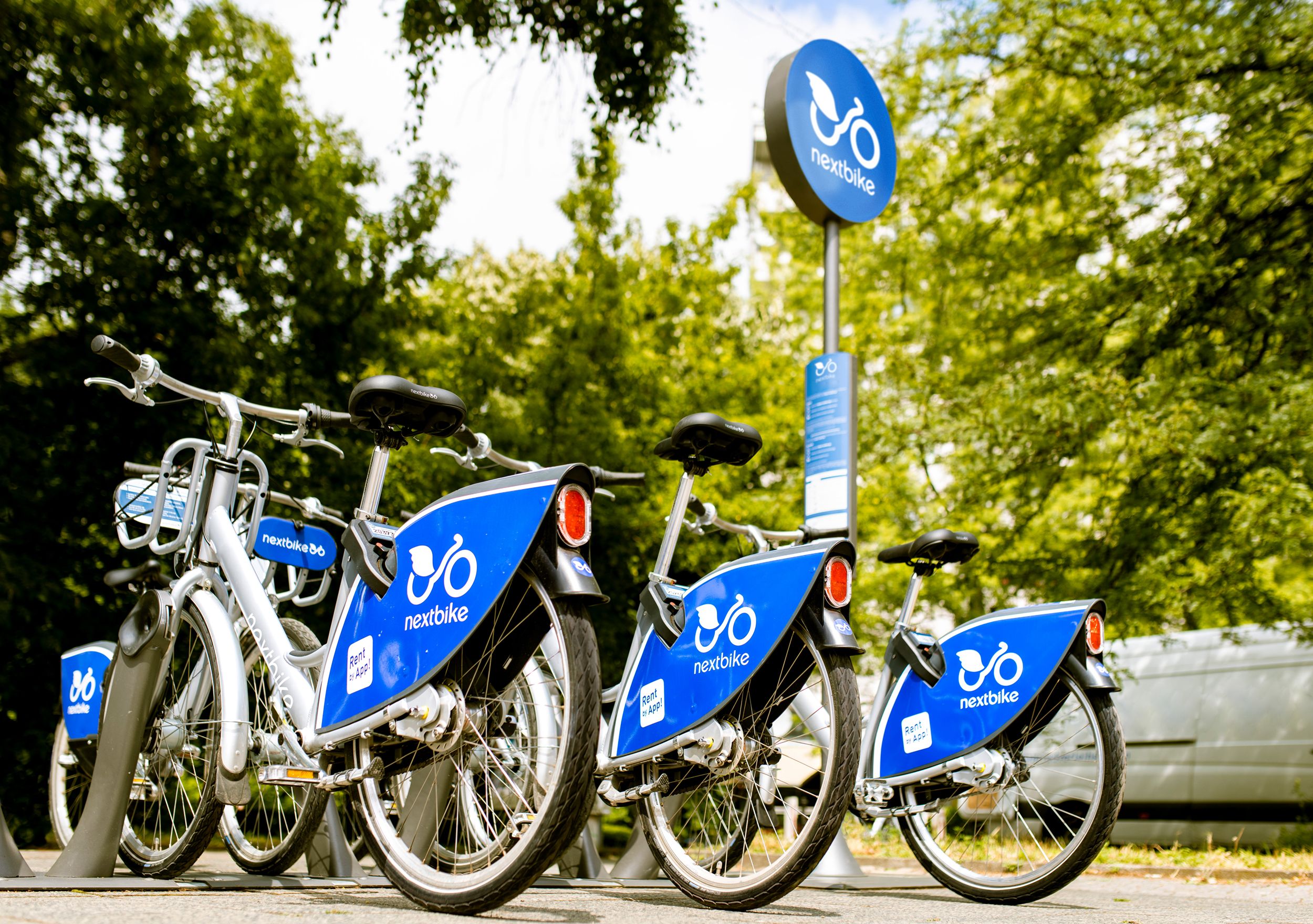 Několik kol Nextbike na venkovní stanici obklopené stromy.