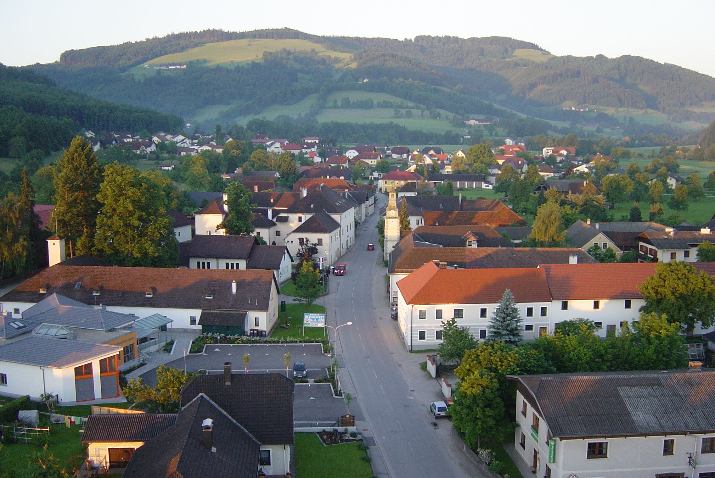 Letecký pohled na vesnici s červenými střechami a zelenou krajinou v pozadí.