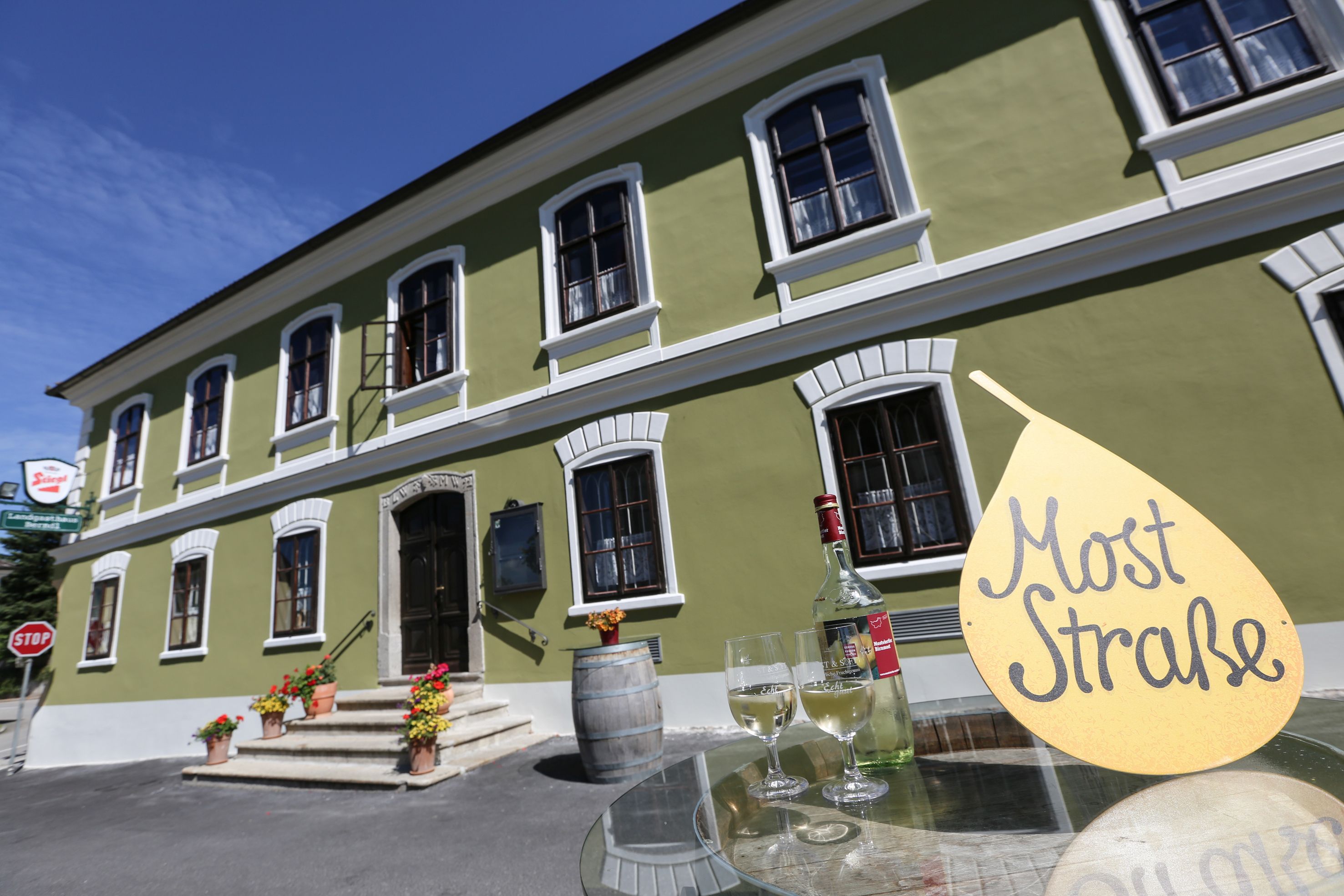 Zelená budova s bílými okny, před ní stůl s lahví vína a sklenicemi, cedule s nápisem "Most Straße".
