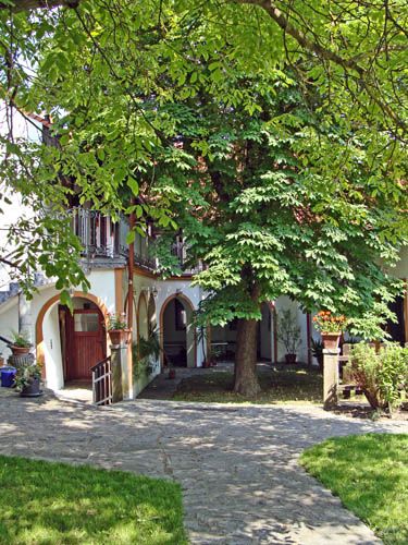 Idylické nádvoří s velkým stromem, obklopené budovou s arkádami a balkony.