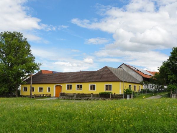 Žlutý statek na zelené louce s modrou oblohou a mraky.
