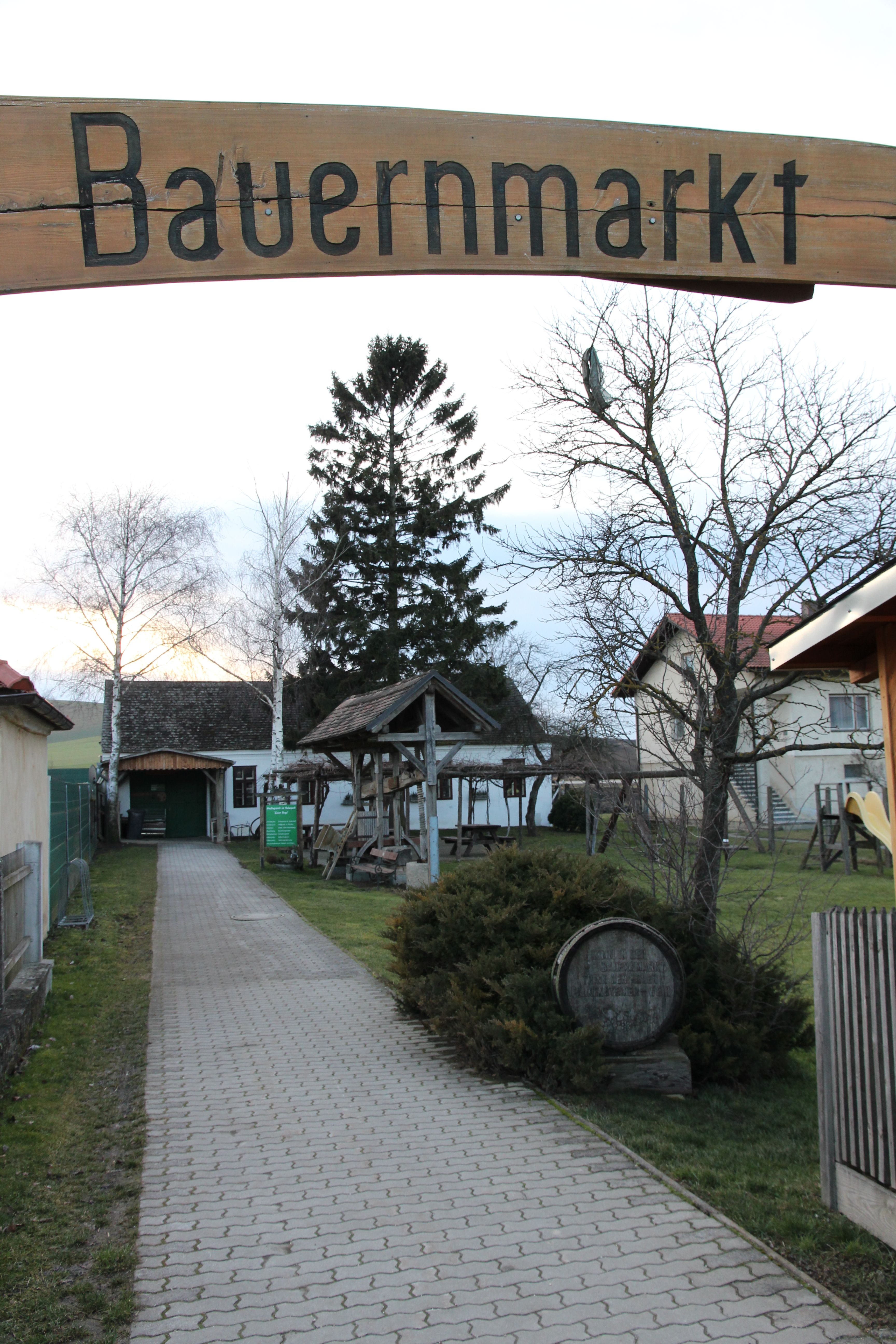 Vchod na farmářský trh s dřevěnou cedulí a dlážděnou cestou obklopenou stromy a budovami.