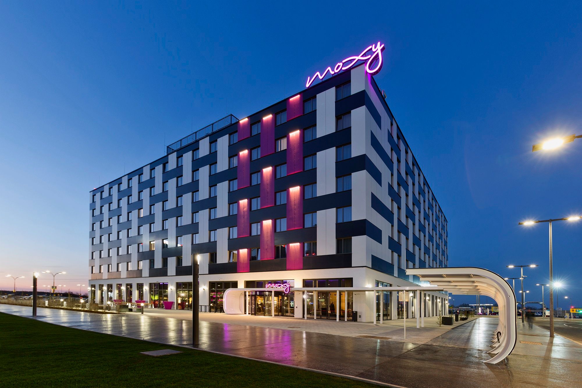Pohled na exteriér hotelu Moxy Vienna Airport za soumraku s osvětlenými nápisy.