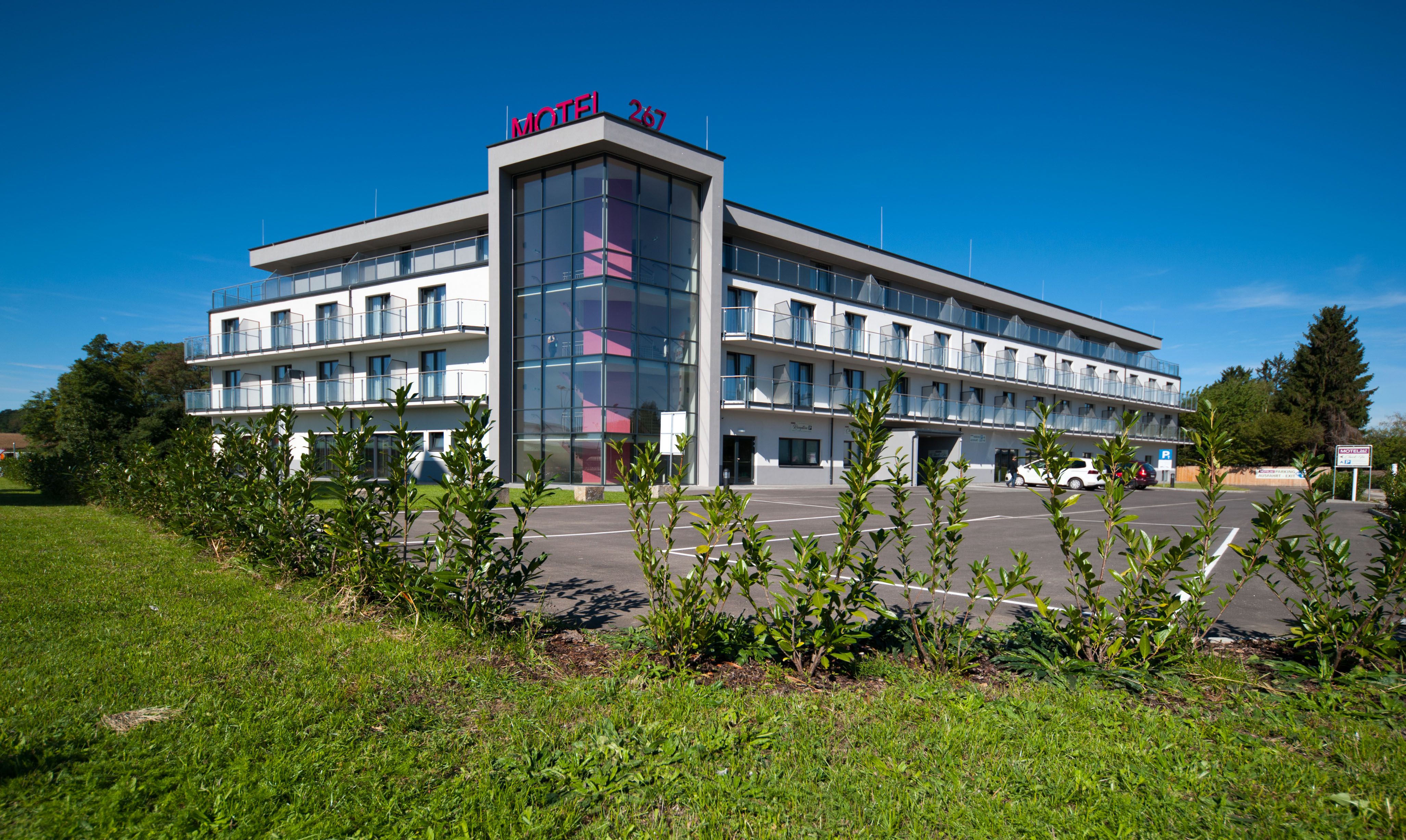Moderní třípodlažní motel s prosklenou fasádou a parkovištěm, obklopený zelení.