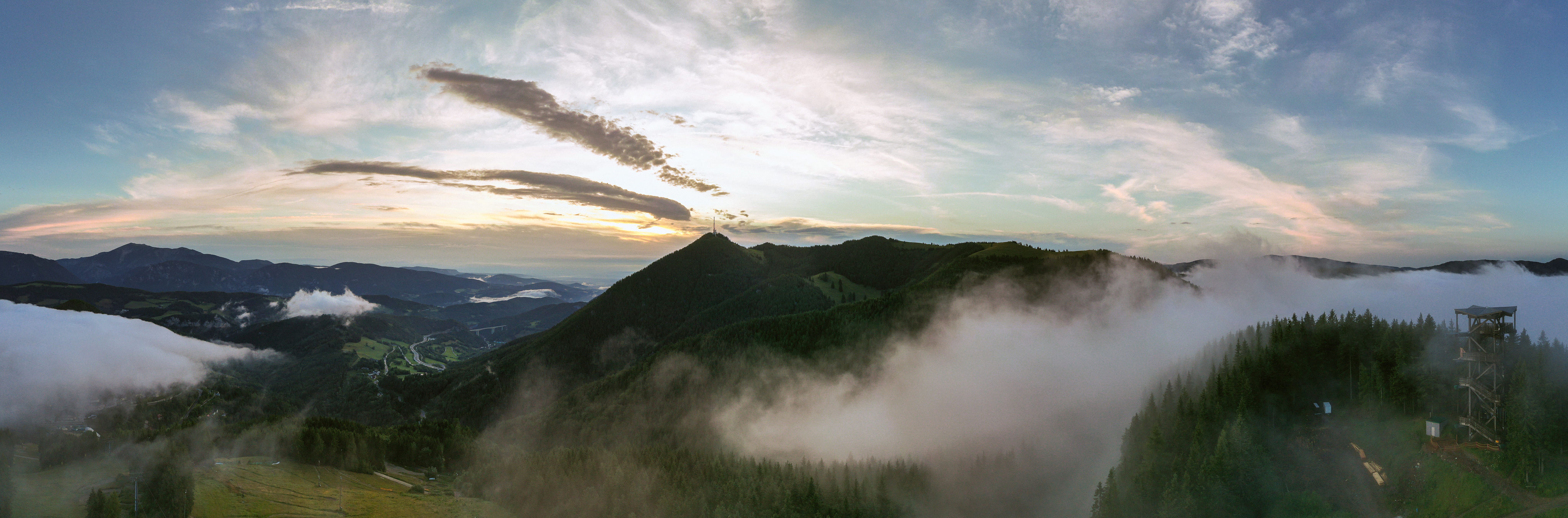Panoramatický pohled na Sonnwendstein při východu slunce s mlhou a rozhlednou v popředí.