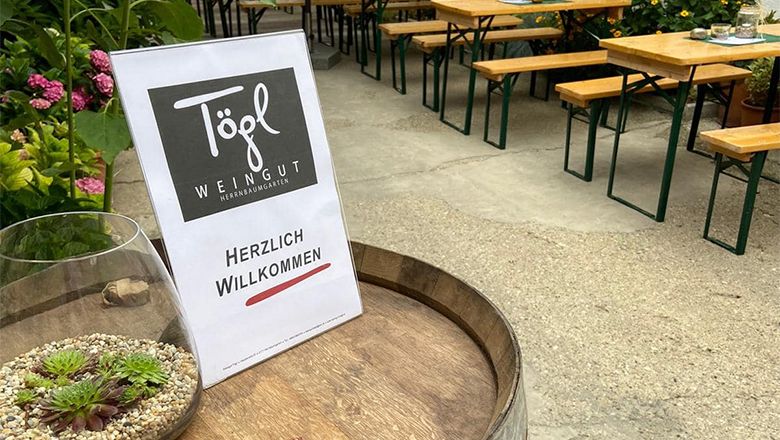 Na sudu s vínem je nápis "Weingut Tögl" a "Herzlich Willkommen" (Vítejte). V pozadí jsou vidět pivní stoly a rostliny.