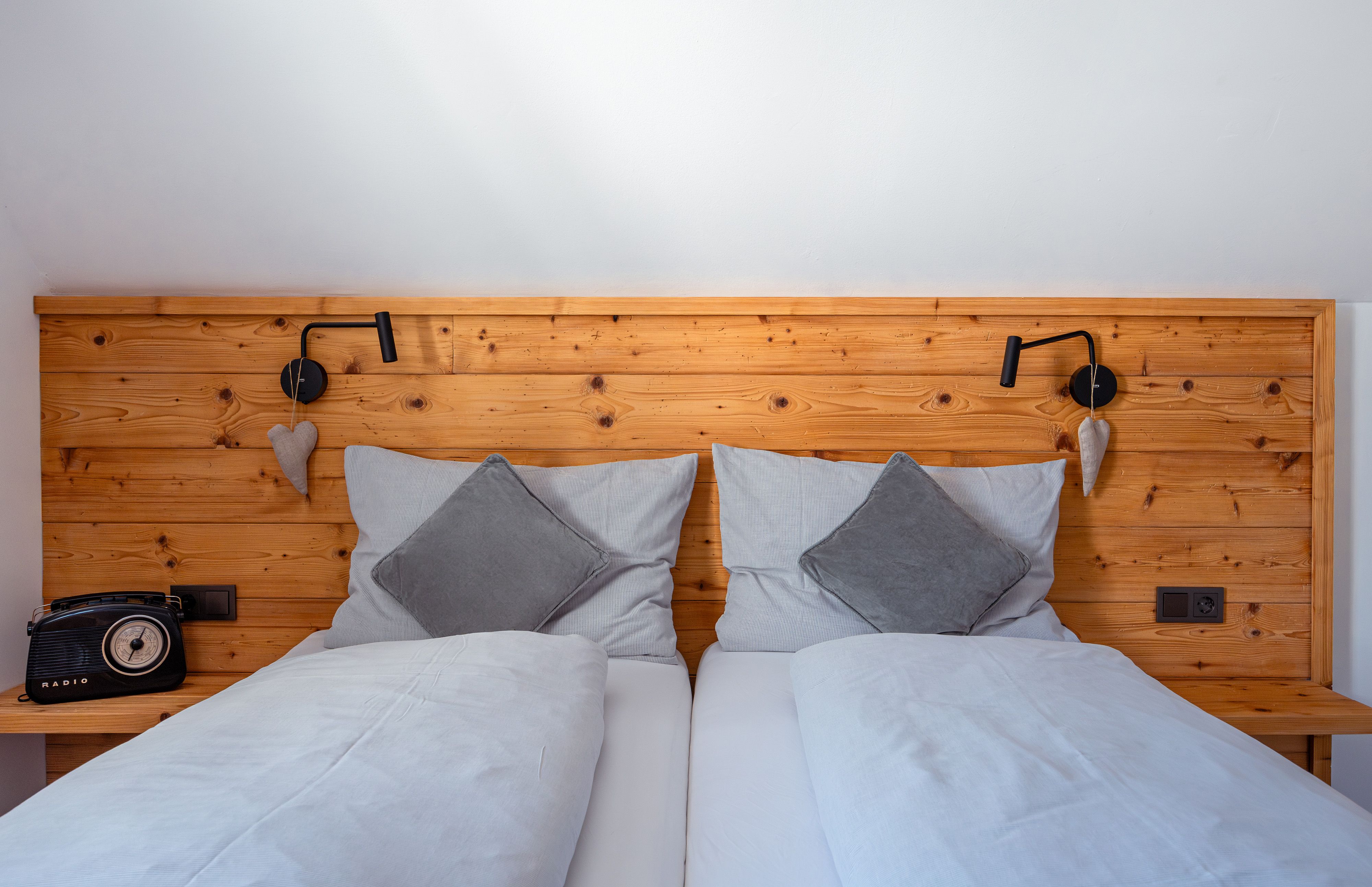 Útulná manželská postel s dřevěným opěradlem, šedými polštáři a přikrývkami. Vlevo retro rádio, vpravo lampa na čtení a dekorace srdce.