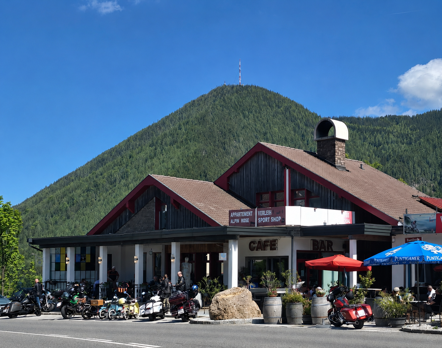 Kavárna a bar s motorkami před hotelem, obklopené horami a stromy.