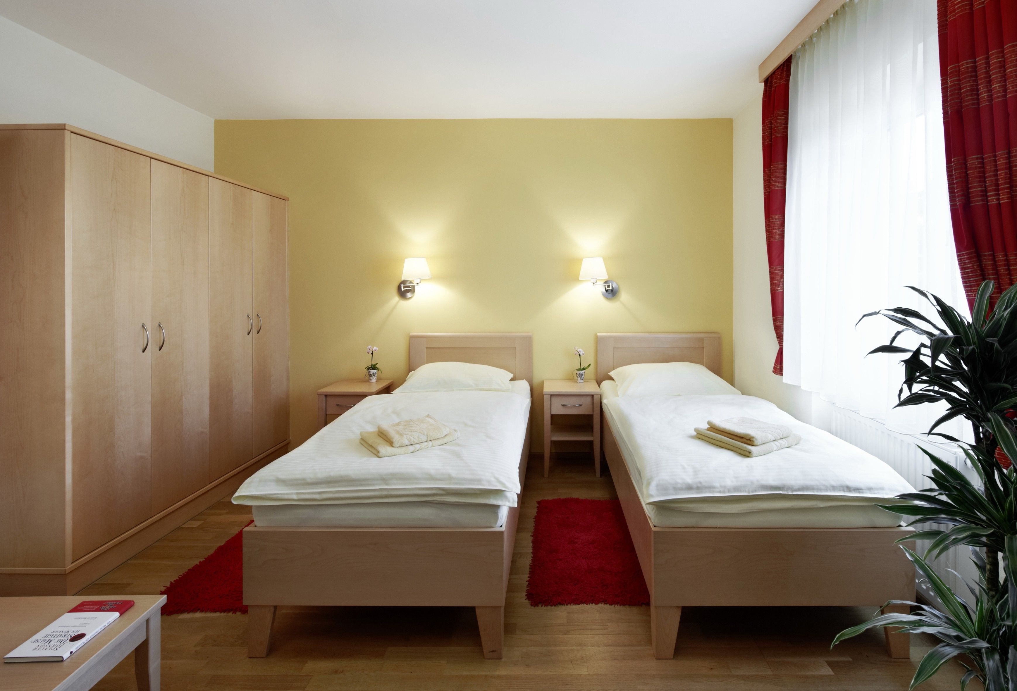 Pokoj pro hosty se dvěma samostatnými lůžky, velkou šatní skříní, červenými závěsy a rostlinou.