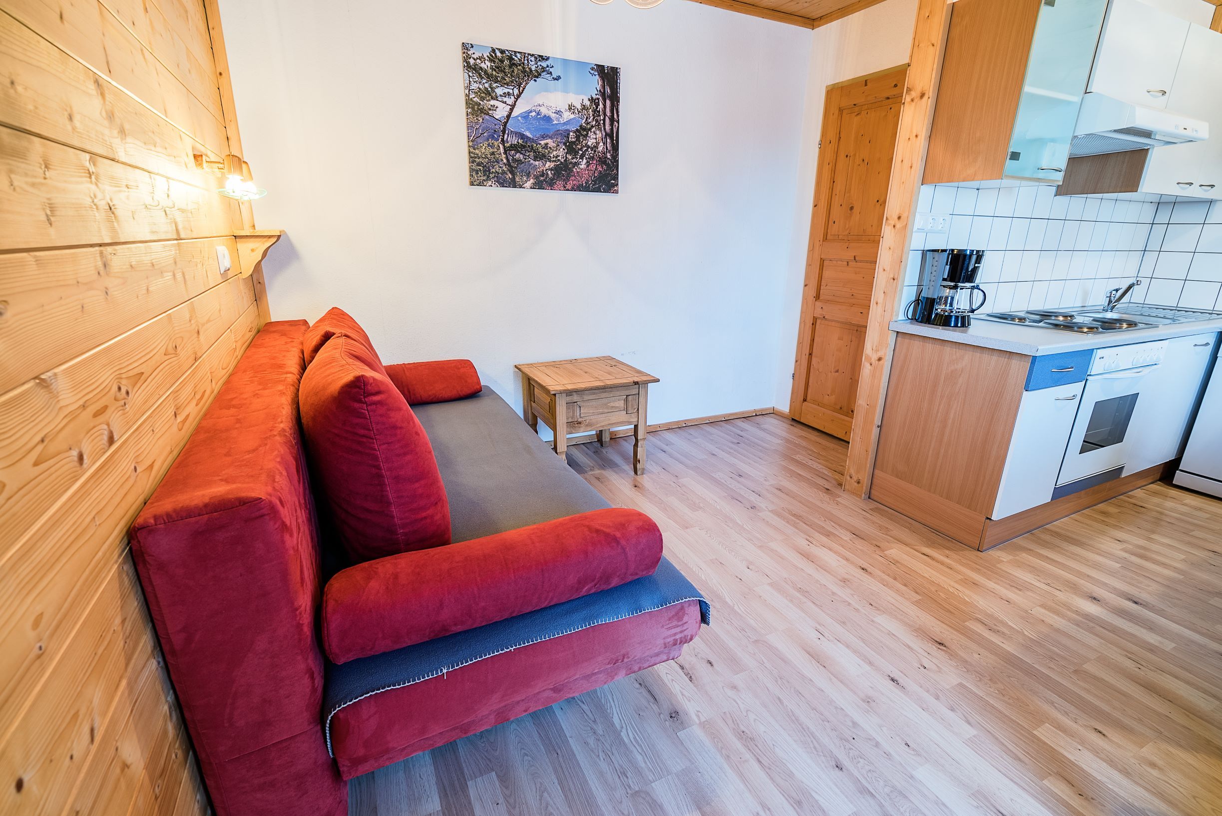 Obývací pokoj s červenou pohovkou, dřevěnými stěnami a malou kuchyňkou.