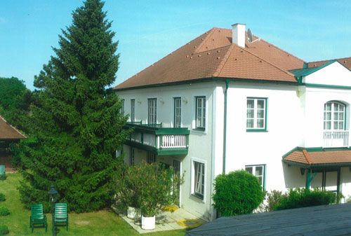 Dvoupodlažní bílý penzion s červenou střechou a zelenými okenicemi, obklopený zahradou se stromy a zahradním nábytkem.