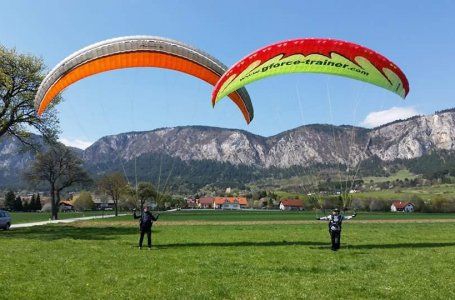 Dva lidé cvičí paragliding na louce před horskou krajinou.
