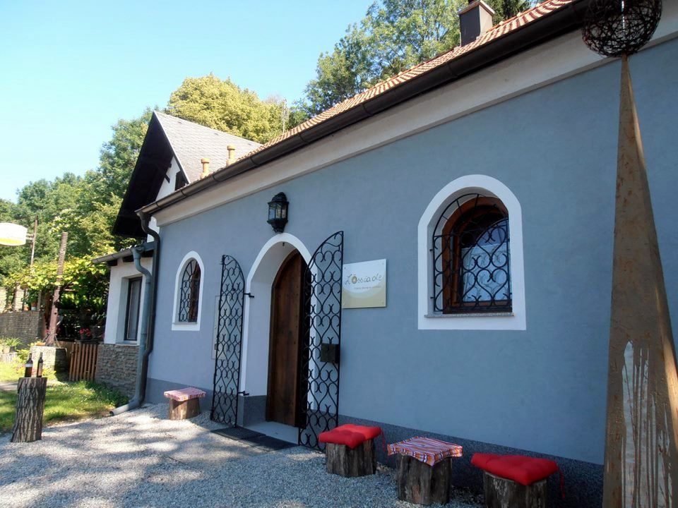 Malá modrá budova s dřevěnými dveřmi a ozdobnými mřížemi před okny. Před ní stojí dřevěné stoličky s červenými polštáři.