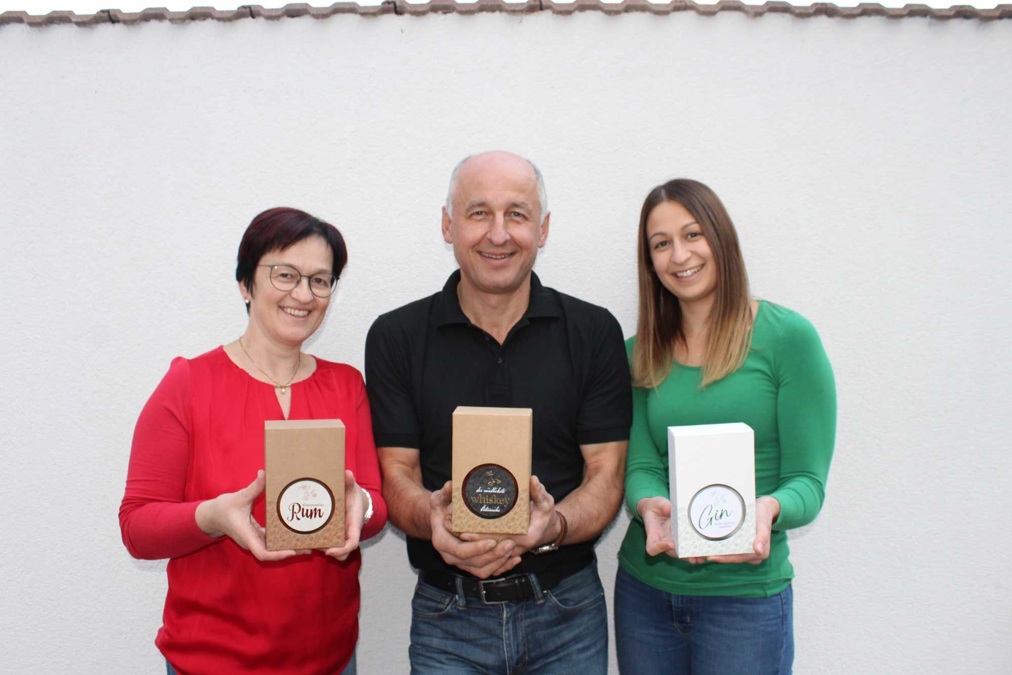 Tři lidé drží před bílou stěnou dárkové krabice s rumem, whisky a ginem.