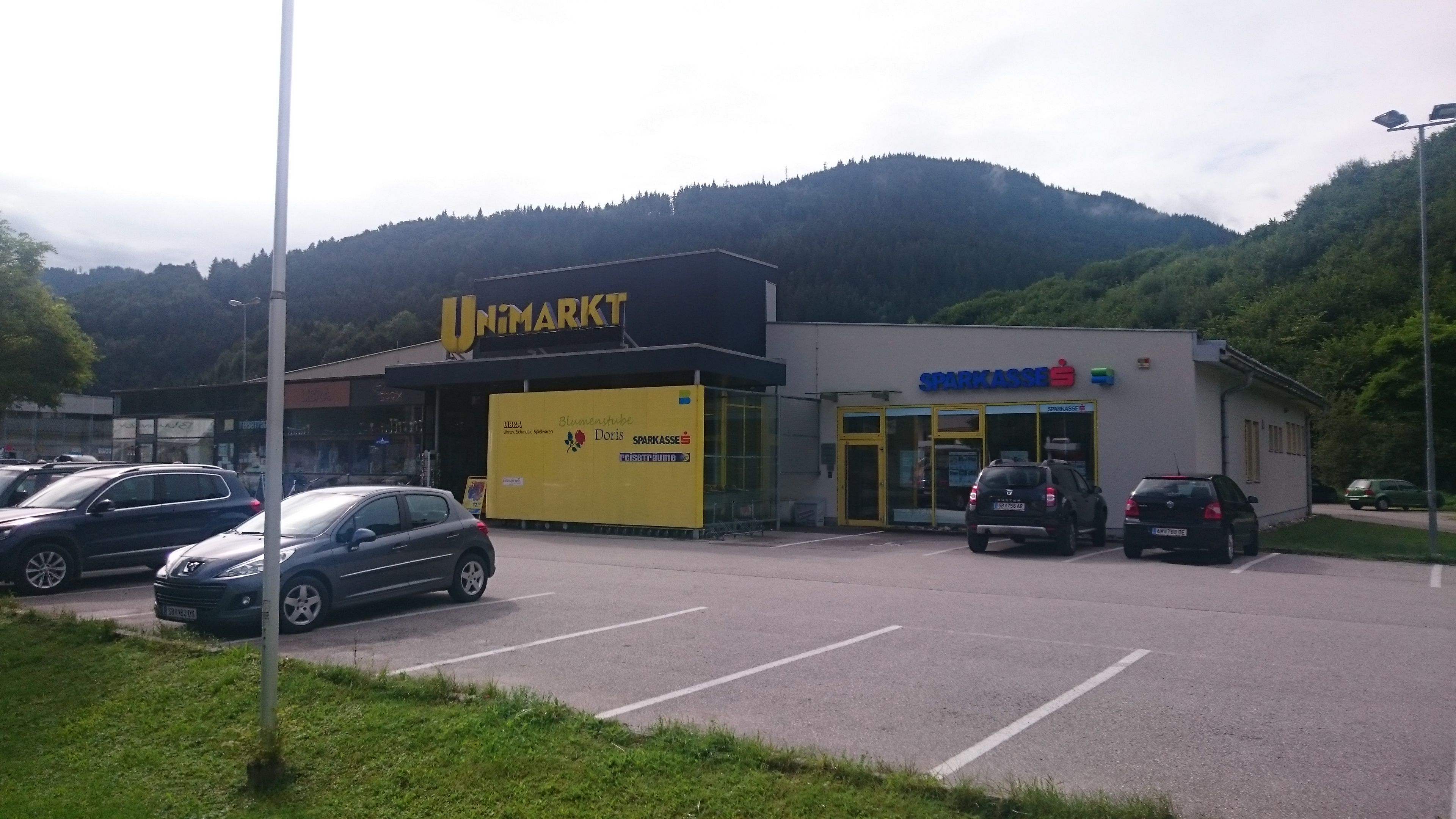 Nákupní centrum se supermarketem Unimarkt a spořitelnou před zalesněnými kopci.